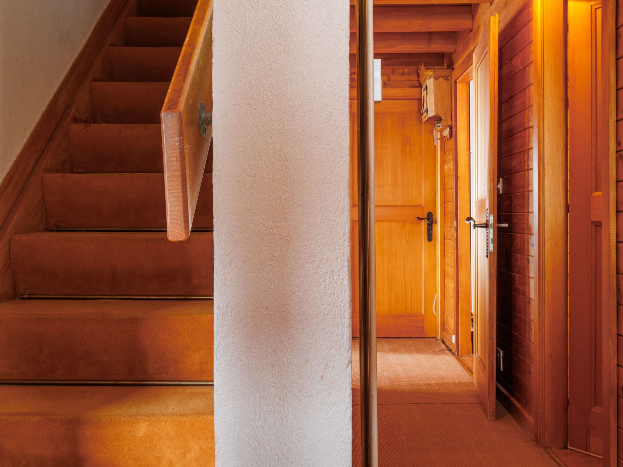 Un escalier en bois mène à un couloir doté de portes en bois et dont le sol est recouvert d’une moquette.