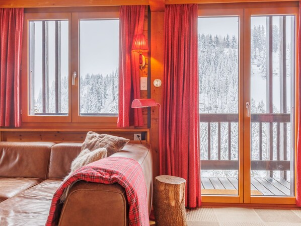 A cozy cabin room with a wooden interior, large windows, a red lamp, and a view of a snowy forest.