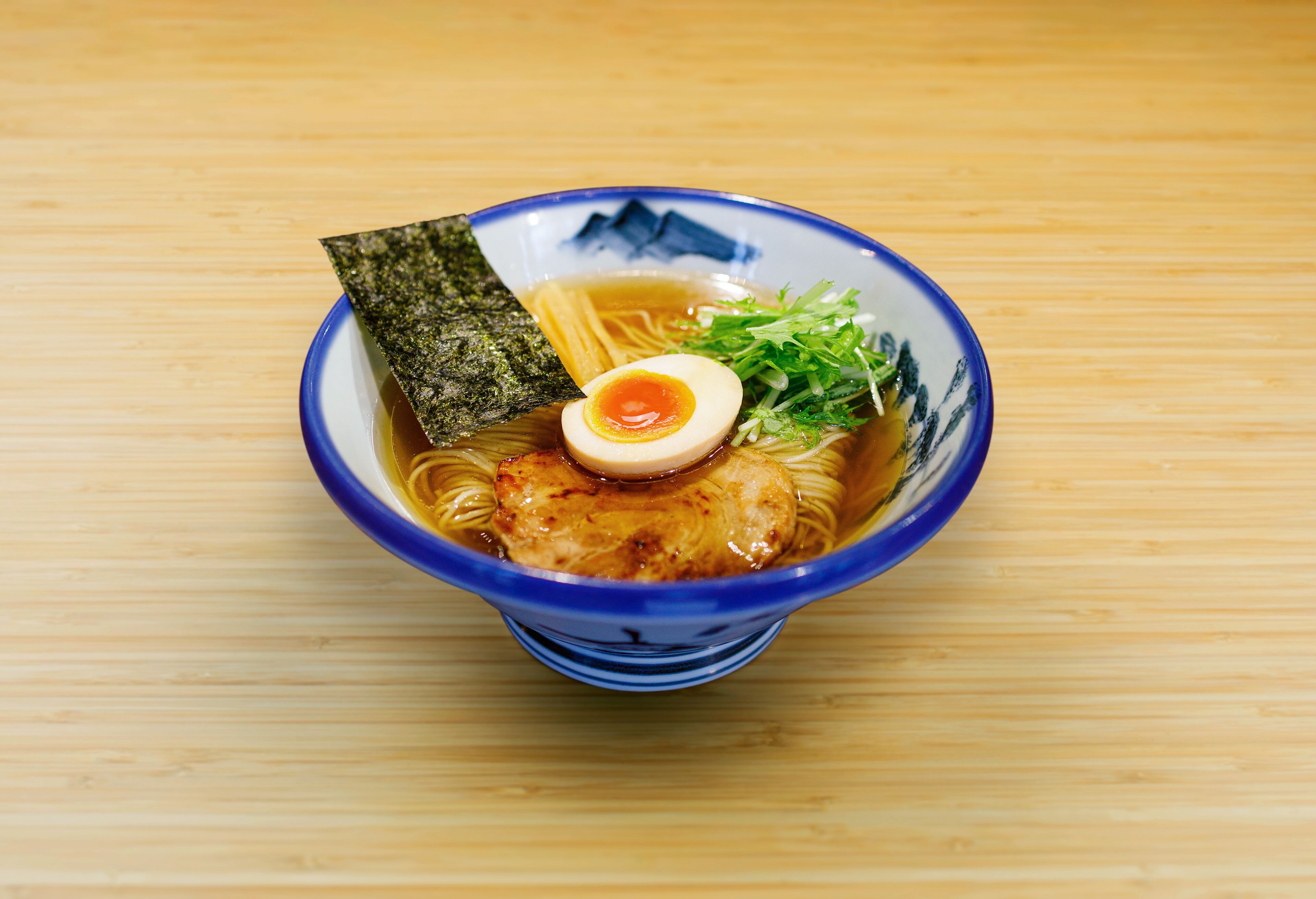 Un tazón de ramen con fideos, una porción de carne, un huevo duro, alga marina y guarnición verde sobre una mesa de madera.