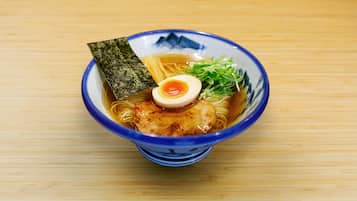 Un tazón de ramen con fideos, una porción de carne, un huevo duro, alga marina y guarnición verde sobre una mesa de madera.