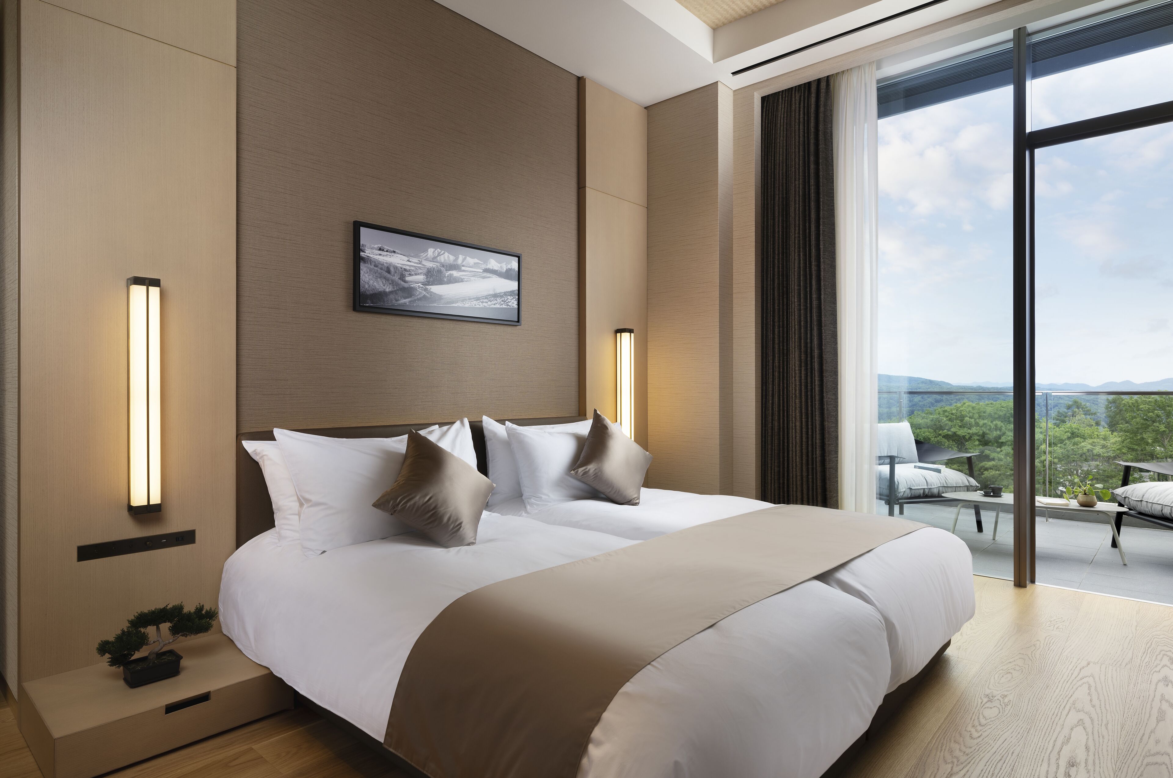 A modern hotel room with a large bed, a view of the outdoors, and a small balcony with seating.