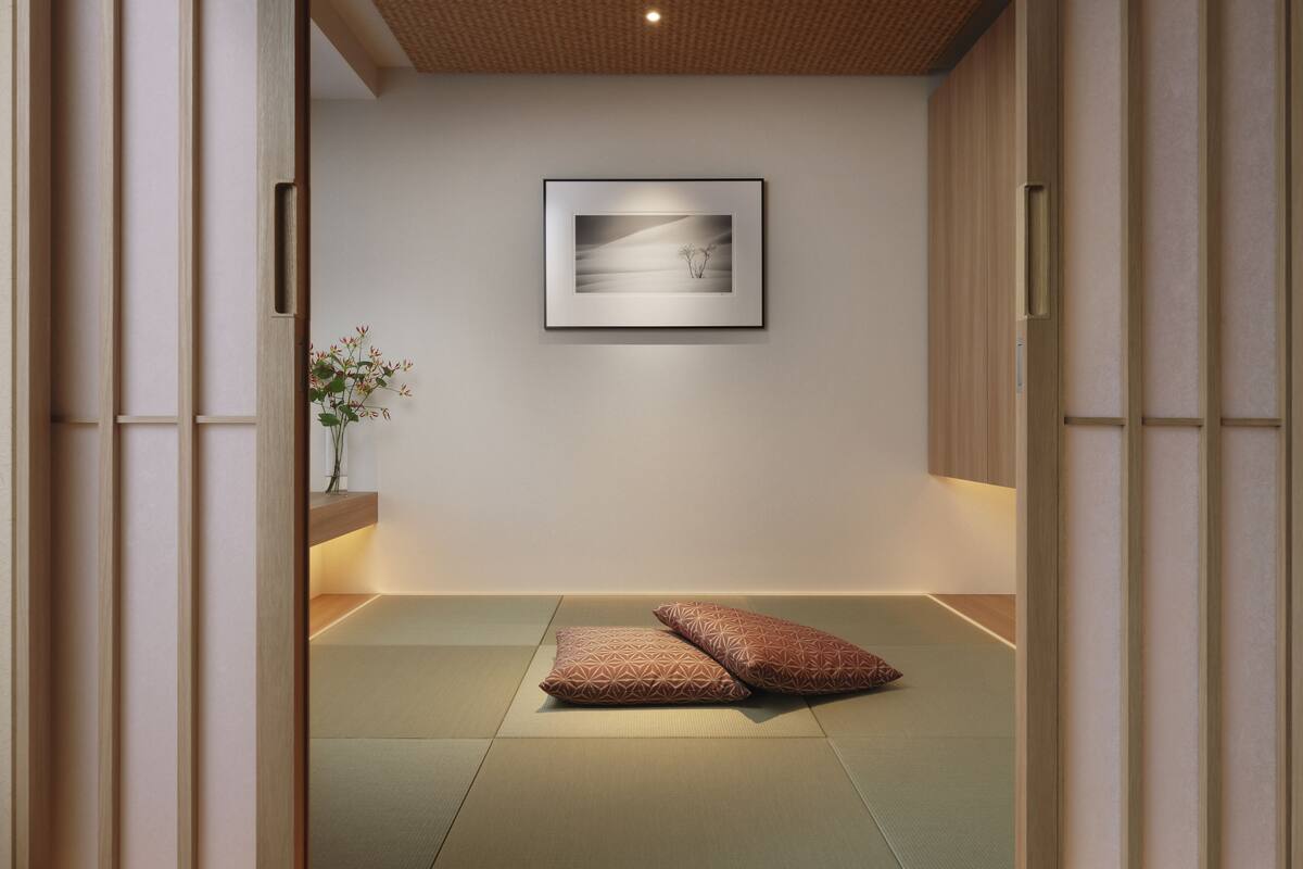 A tatami mat room with a framed picture on the wall, a small vase with flowers, and two pillows.