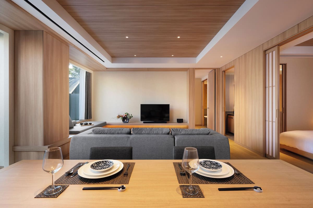 A modern hotel room with a wooden ceiling, a large sofa, a TV, and a dining table set with plates and glasses.