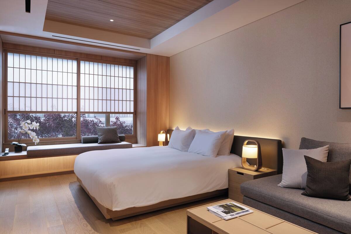 A hotel room with a bed, a sofa, a nightstand, a lamp, a framed picture on the wall, and a window with a view of trees.