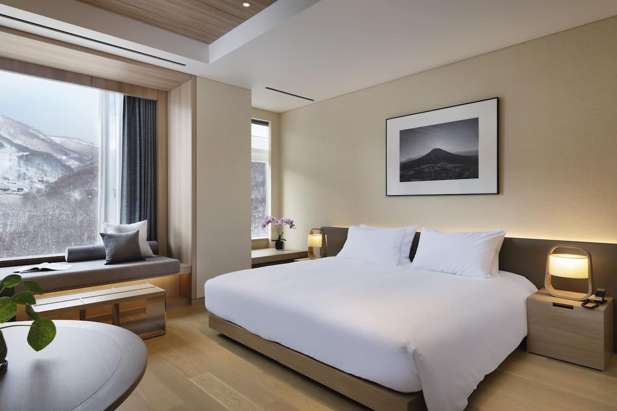 A modern hotel room with a large bed, a seating area by the window, a bedside table with a lamp, and a framed picture on the wall.