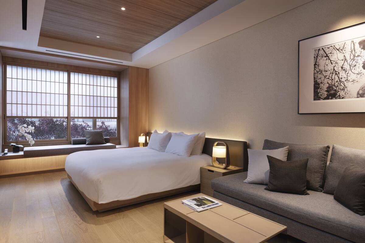 A modern hotel room with a large bed, a sofa, a bedside table with a lamp, and a window with a view of trees.