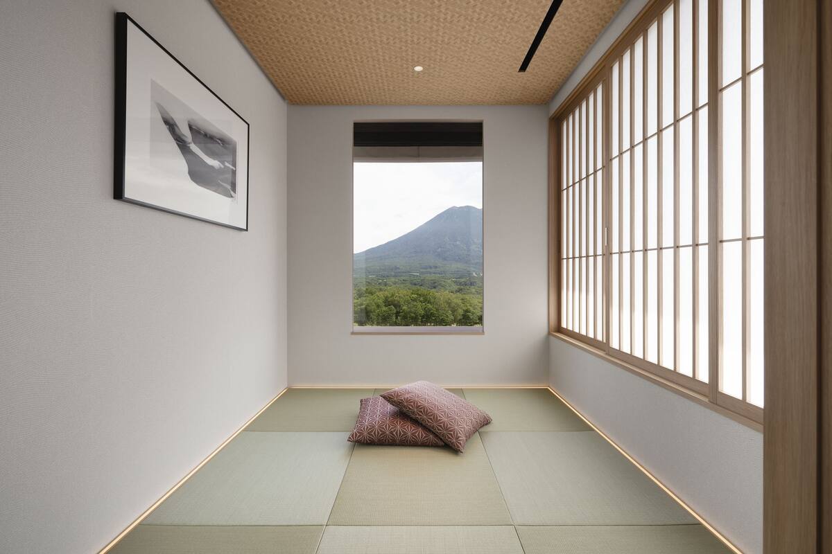 A minimalist room with tatami flooring, a framed picture on the wall, a window with a view of a mountain, and a sliding door with a wooden frame.