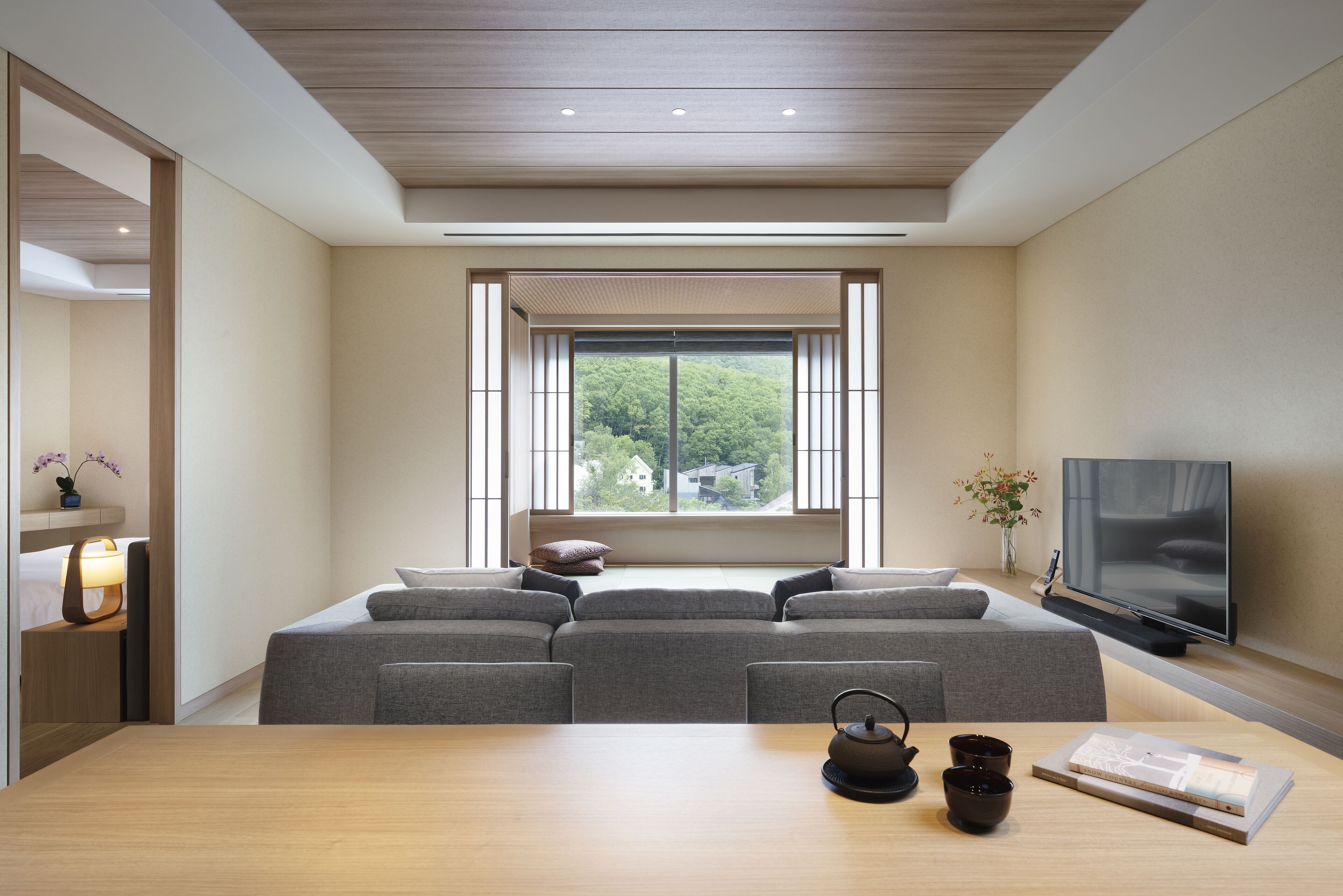 A modern living room with a sofa, a wooden table, a television, and a window with a view of greenery.