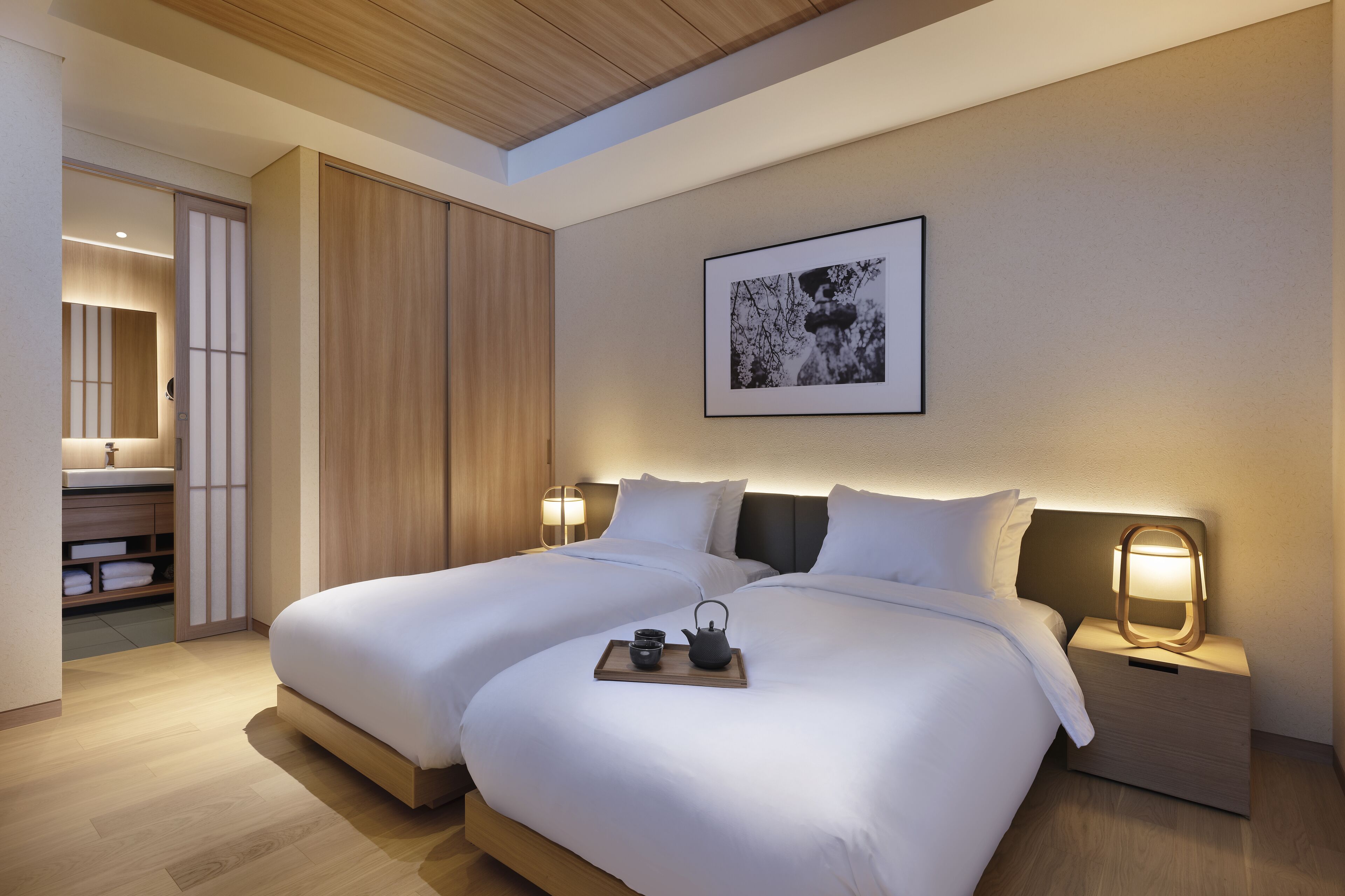 Habitación de hotel moderna con dos camas, una mesita de noche con lámpara, un cuadro enmarcado en la pared y una bandeja con tetera y tazas.