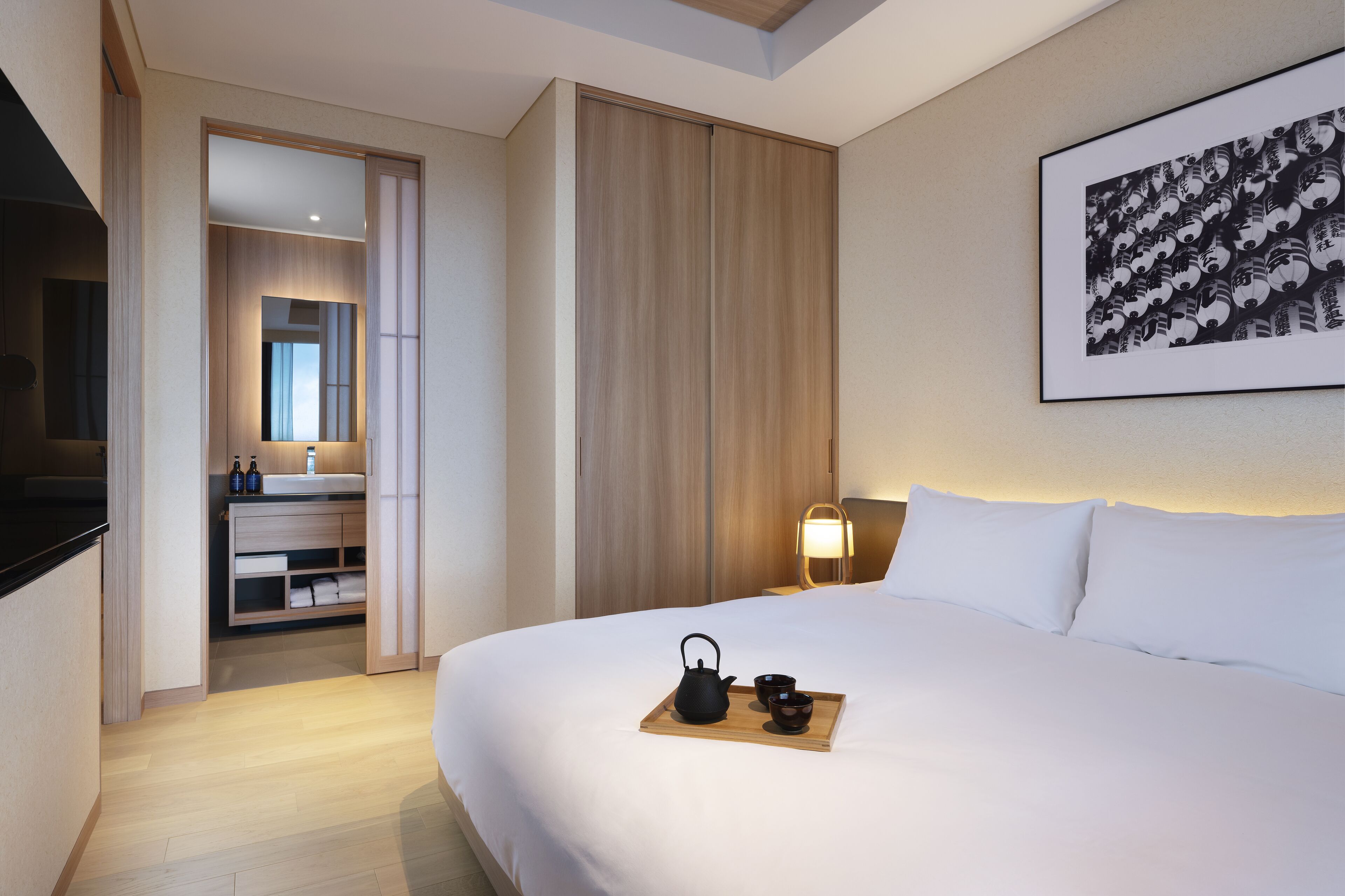 A hotel room with a large bed, a bedside table with a lamp, a wooden side table with a tray of tea, a wardrobe, and a framed picture on the wall.