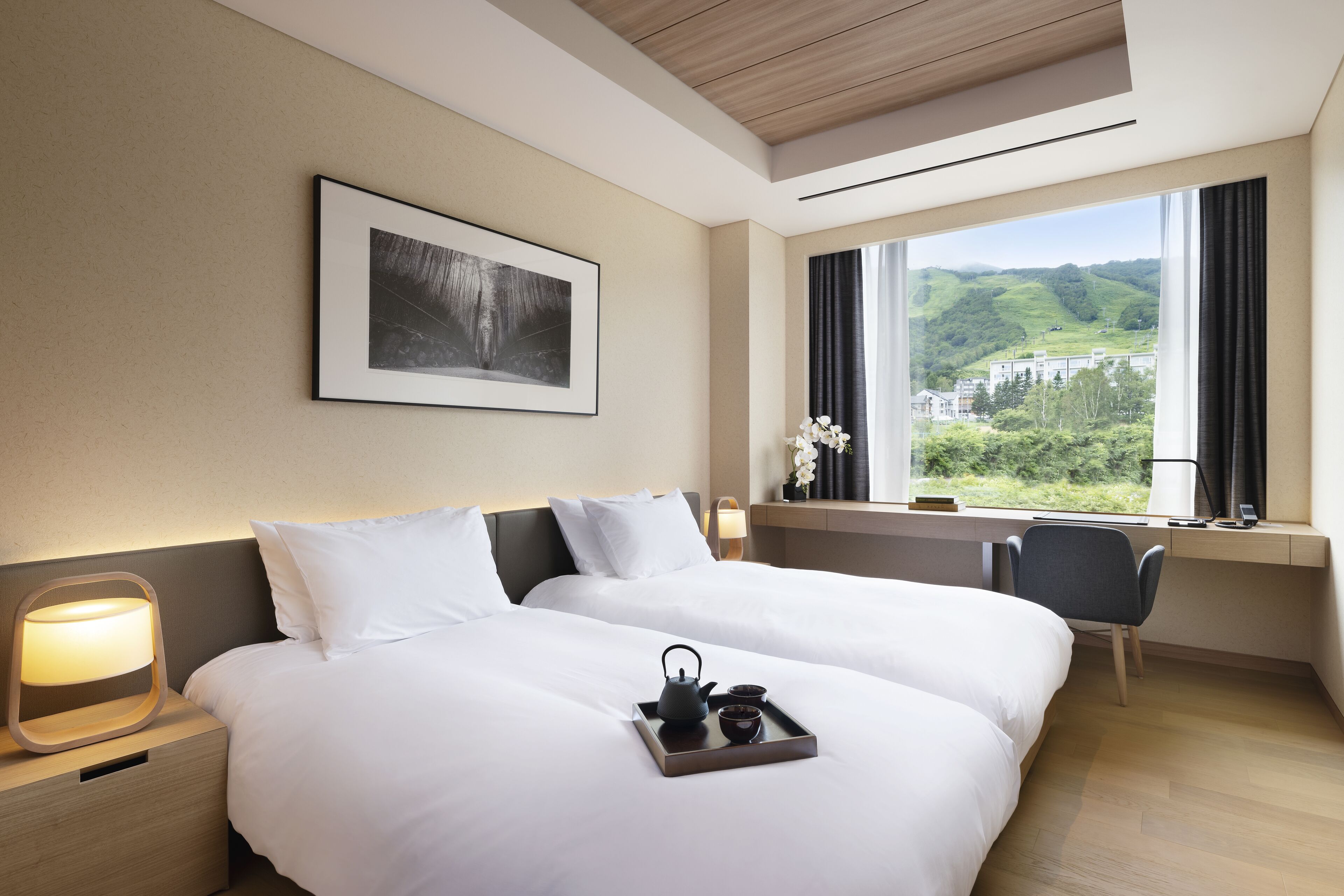 A hotel room with two beds, a desk, a chair, a window with a view of greenery, and a framed picture on the wall.