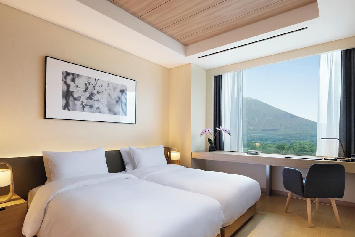 A hotel room with a bed, a desk, a chair, a window with a view of a mountain, and a framed picture on the wall.