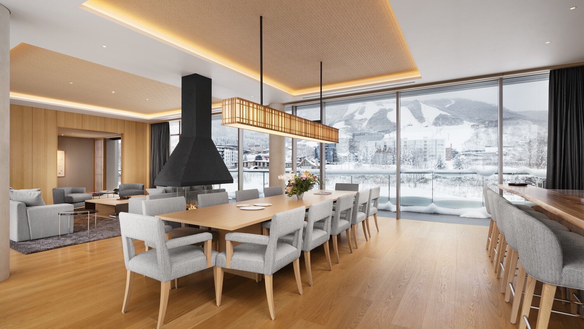 A modern hotel room with a large dining table, comfortable seating, a fireplace, and a view of a snowy mountain landscape.