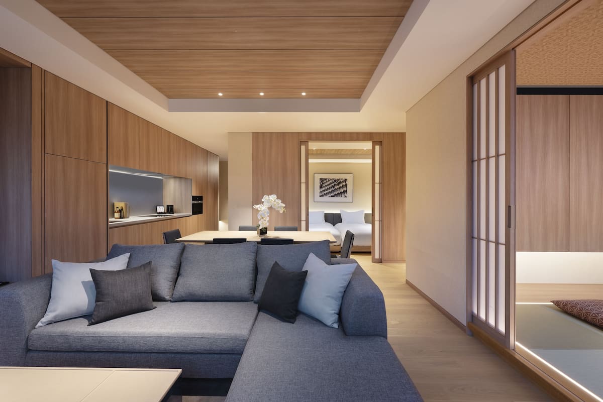 A modern living room with a grey sectional sofa, wooden cabinets, a kitchen area, and a traditional Japanese sliding door.