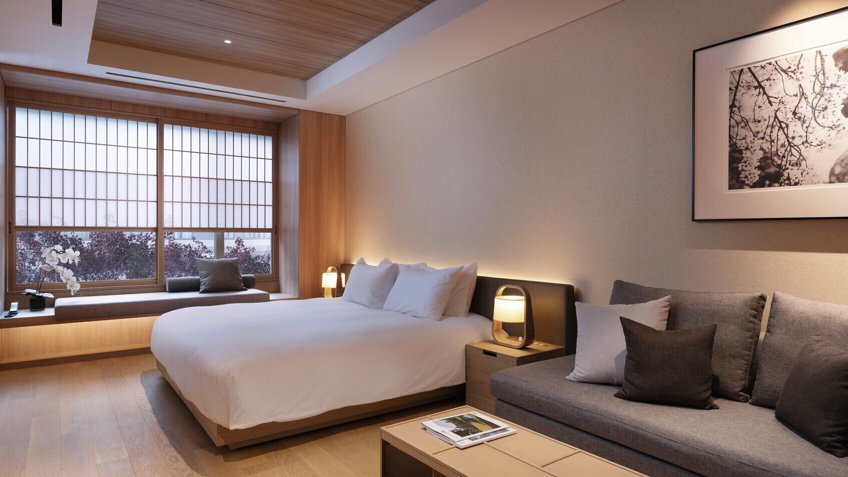 A hotel room with a bed, a sofa, a bedside table, a nightstand, a window with a view, and a framed picture on the wall.