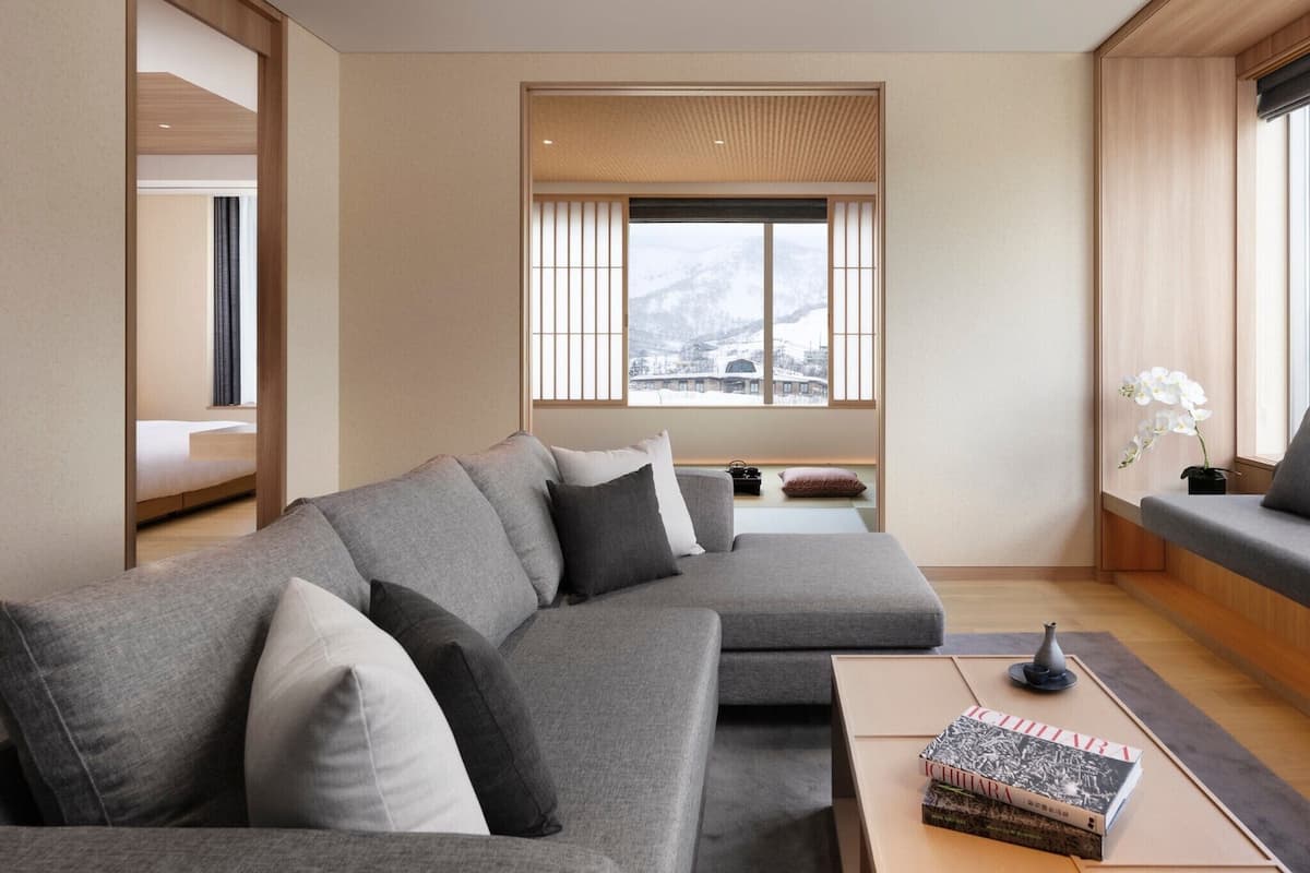 A modern living room with a grey sectional sofa, a small table with books, and a view of snow-capped mountains through large windows.