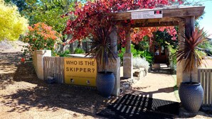 A garden entrance with a wooden pergola, a sign asking "WHO IS THE SKIPPER?", and a signboard for the Producers License. - Vineyard Accomodation - Self Catering (Henty)
