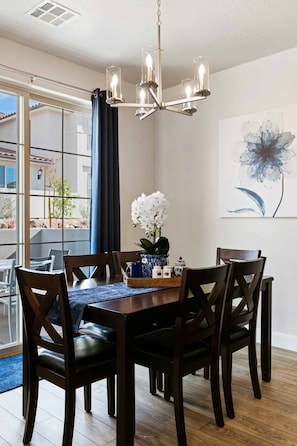 Un comedor con candelabro, sillas de madera, una mesa con un jarrón de flores y un ventanal con vistas a edificios y árboles.
