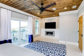 A bedroom with a ceiling fan, a fireplace, a TV mounted on the wall, and a balcony with curtains.