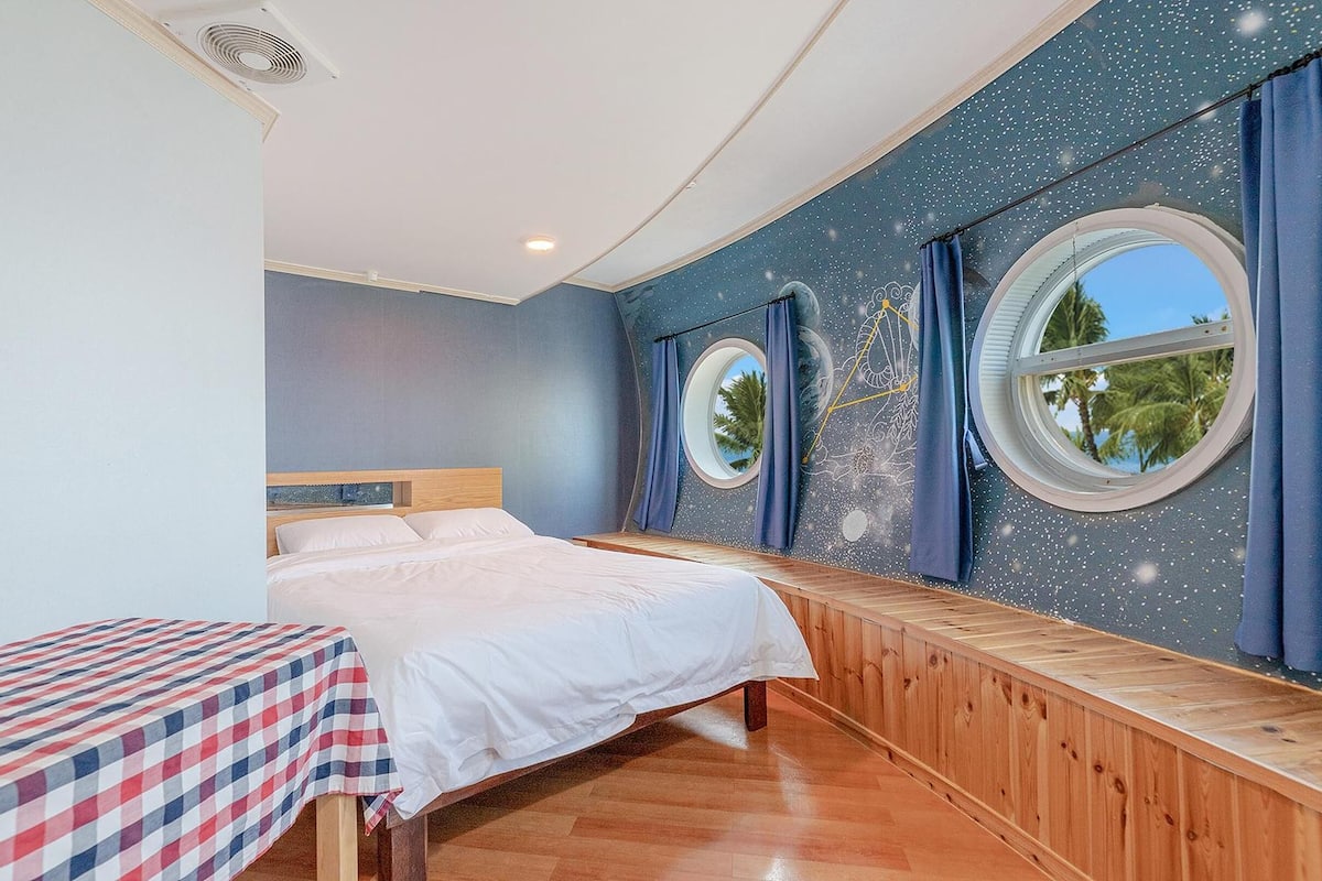 Une chambre avec un lit, un banc, deux fenêtres et une fresque murale représentant un ciel étoilé.