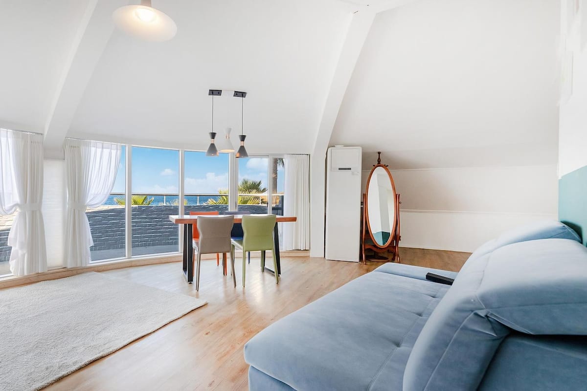 Une pièce spacieuse avec une grande fenêtre donnant sur une plage, un coin repas avec des chaises, un canapé et un miroir.