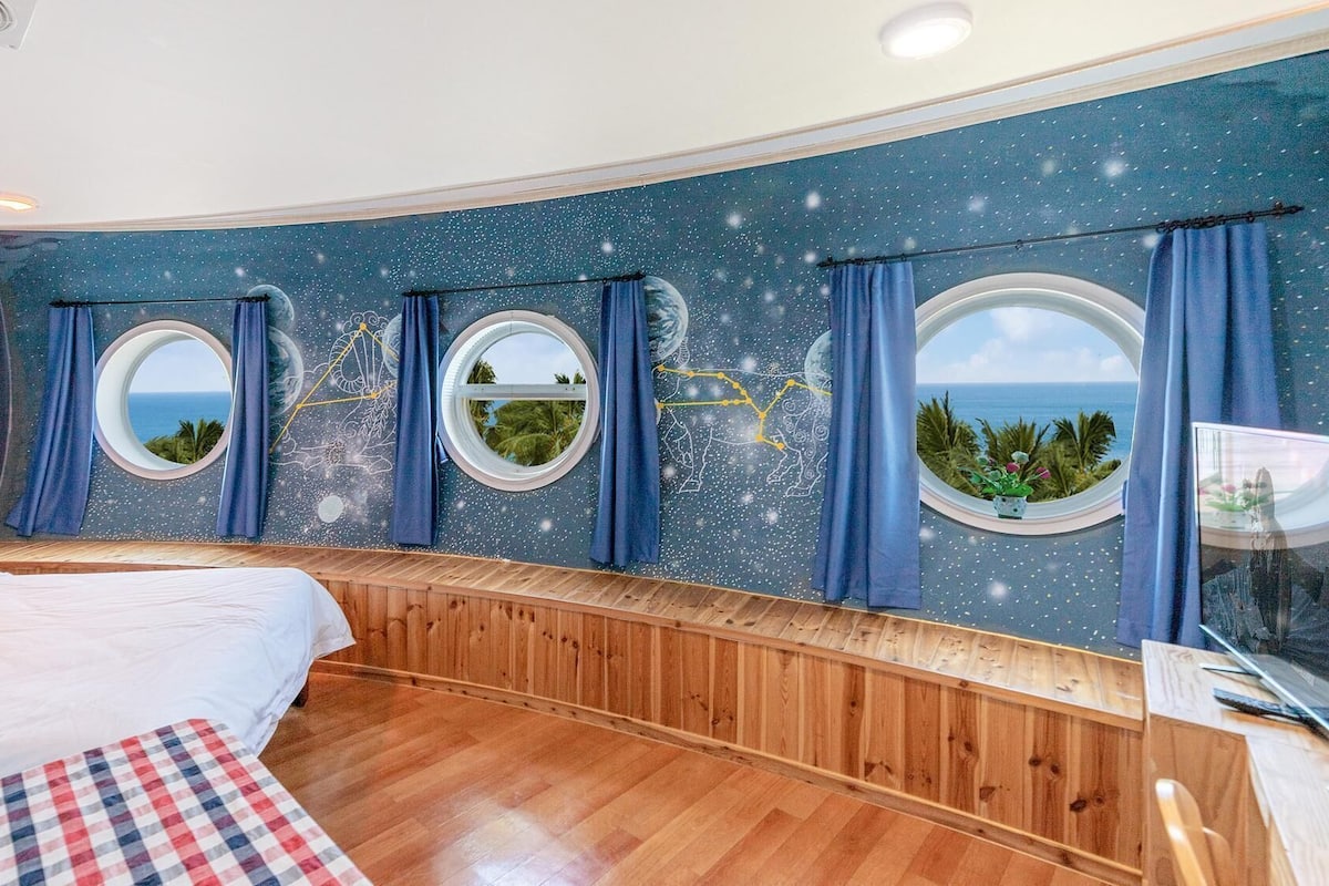 Une chambre à coucher avec un banc en bois aux lignes courbes, un lit et trois fenêtres rondes ornées de rideaux.