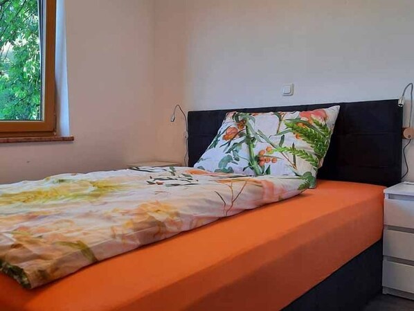 A bed with an orange bedspread and a floral pillow, a nightstand, and a window with a view of trees. - Kiwi apartment with balcony - Kiwi vacation apartment (Weyregg am Attersee)