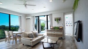 A spacious living area with a sofa, coffee table, dining table, and chairs. There is a ceiling fan, a television mounted on the wall, and a large window offering a view of the outdoors. - Sea Space Road (Onna)