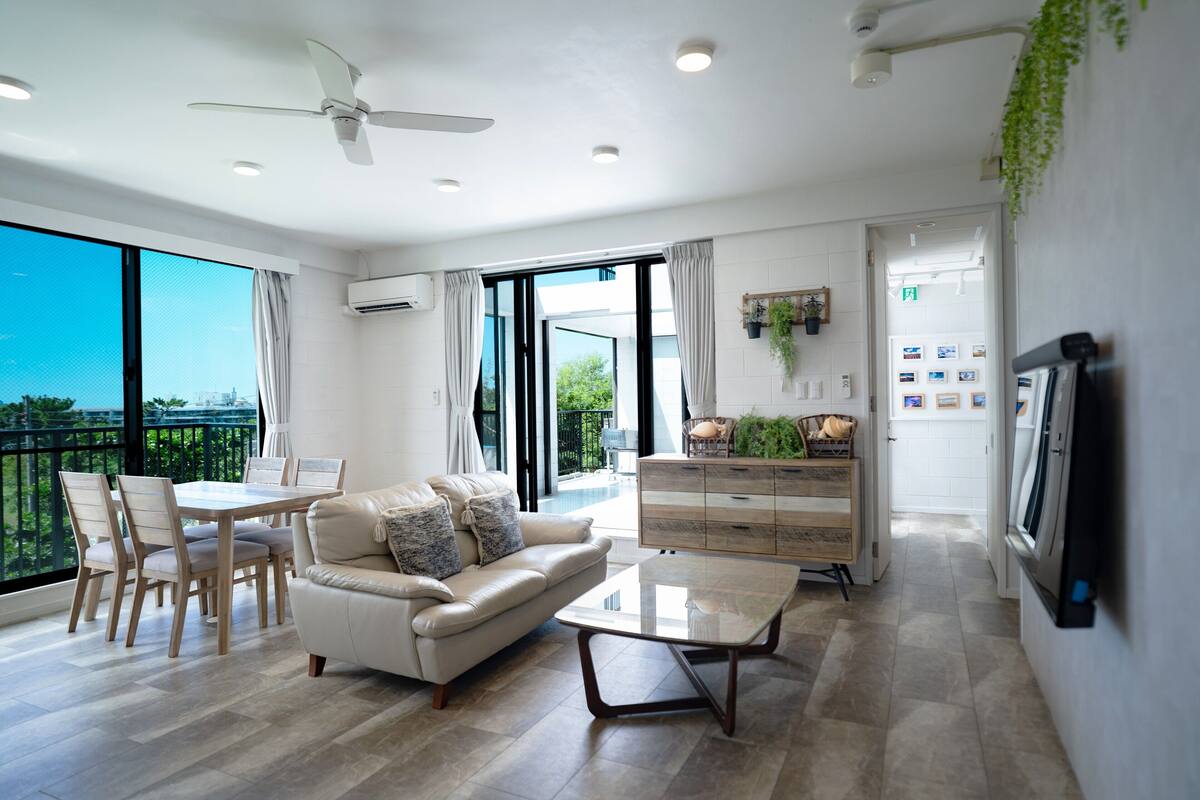 A spacious living area with a sofa, coffee table, dining table, and chairs. There is a ceiling fan, a television mounted on the wall, and a large window offering a view of the outdoors.
