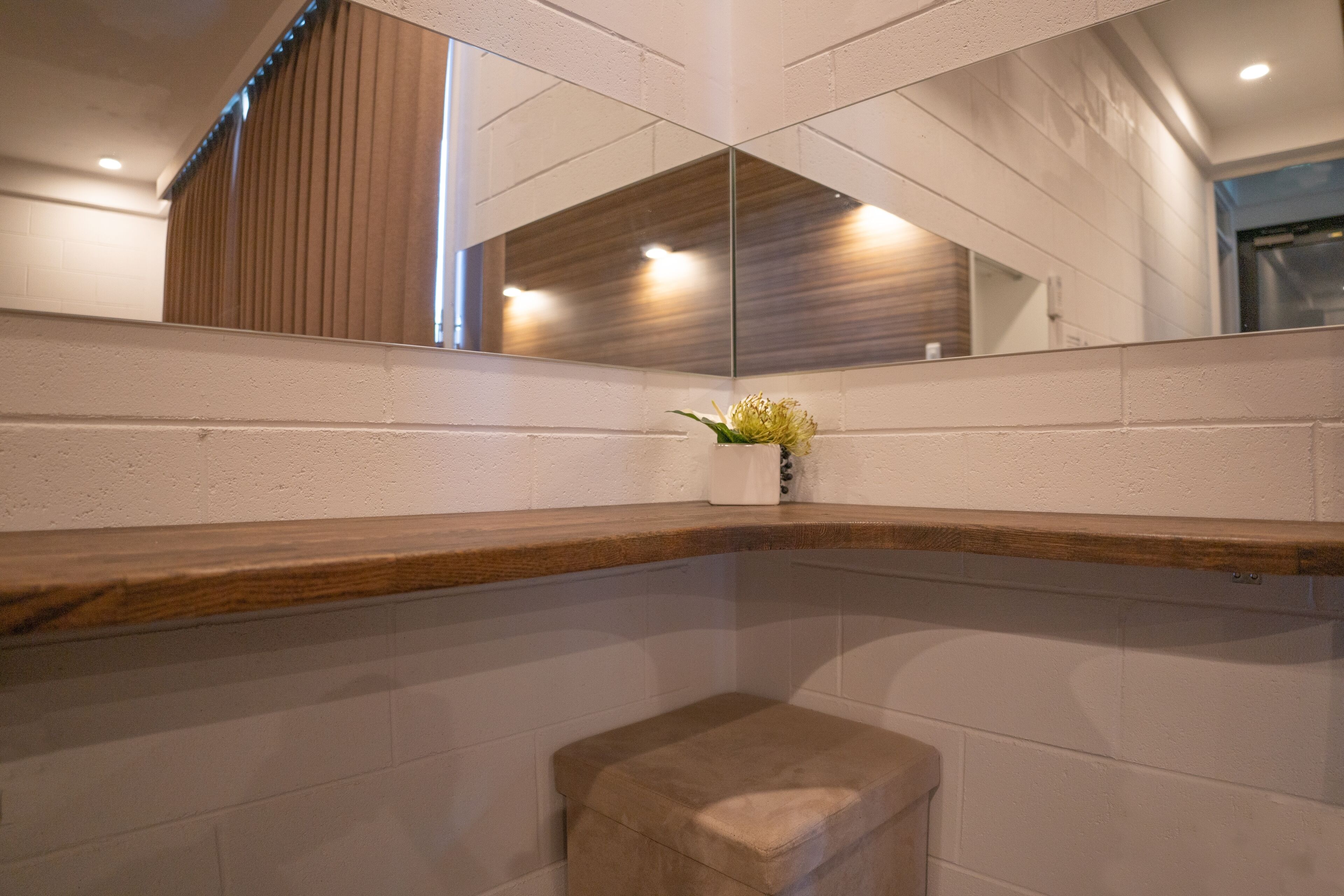 Un baño con tocador de madera, un taburete pequeño y un espejo que refleja un panel de madera y una ventana.