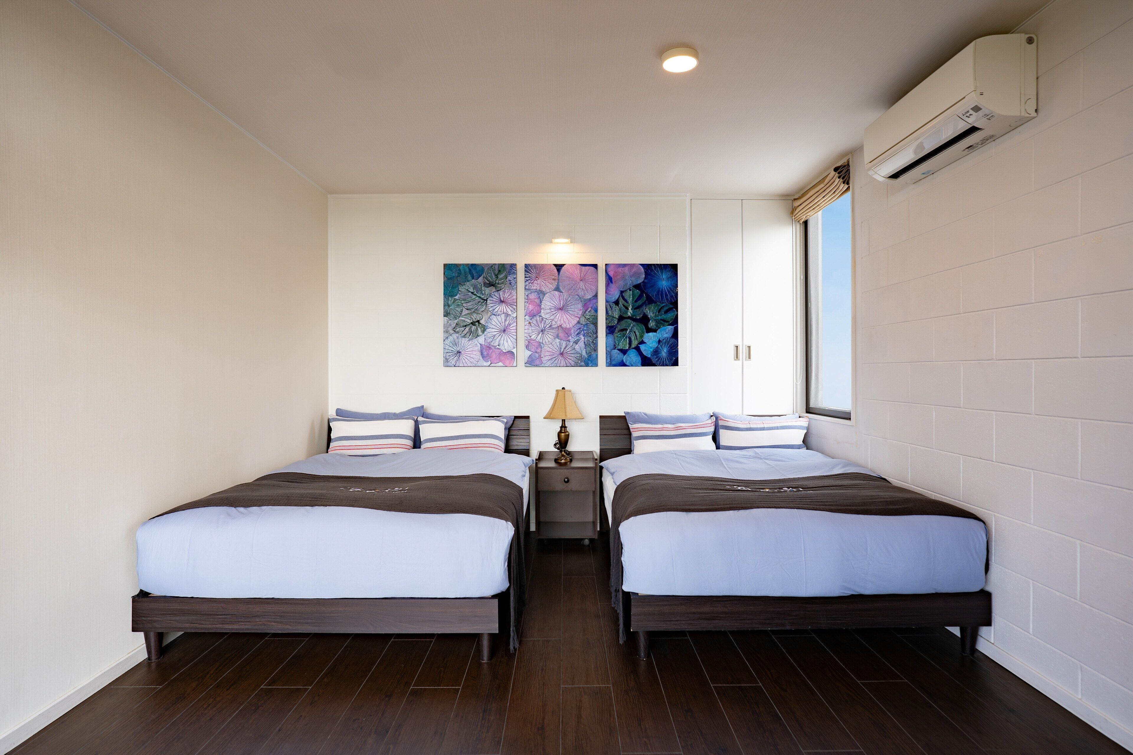 A hotel room with two beds, a nightstand, a lamp, and a wall-mounted air conditioner.