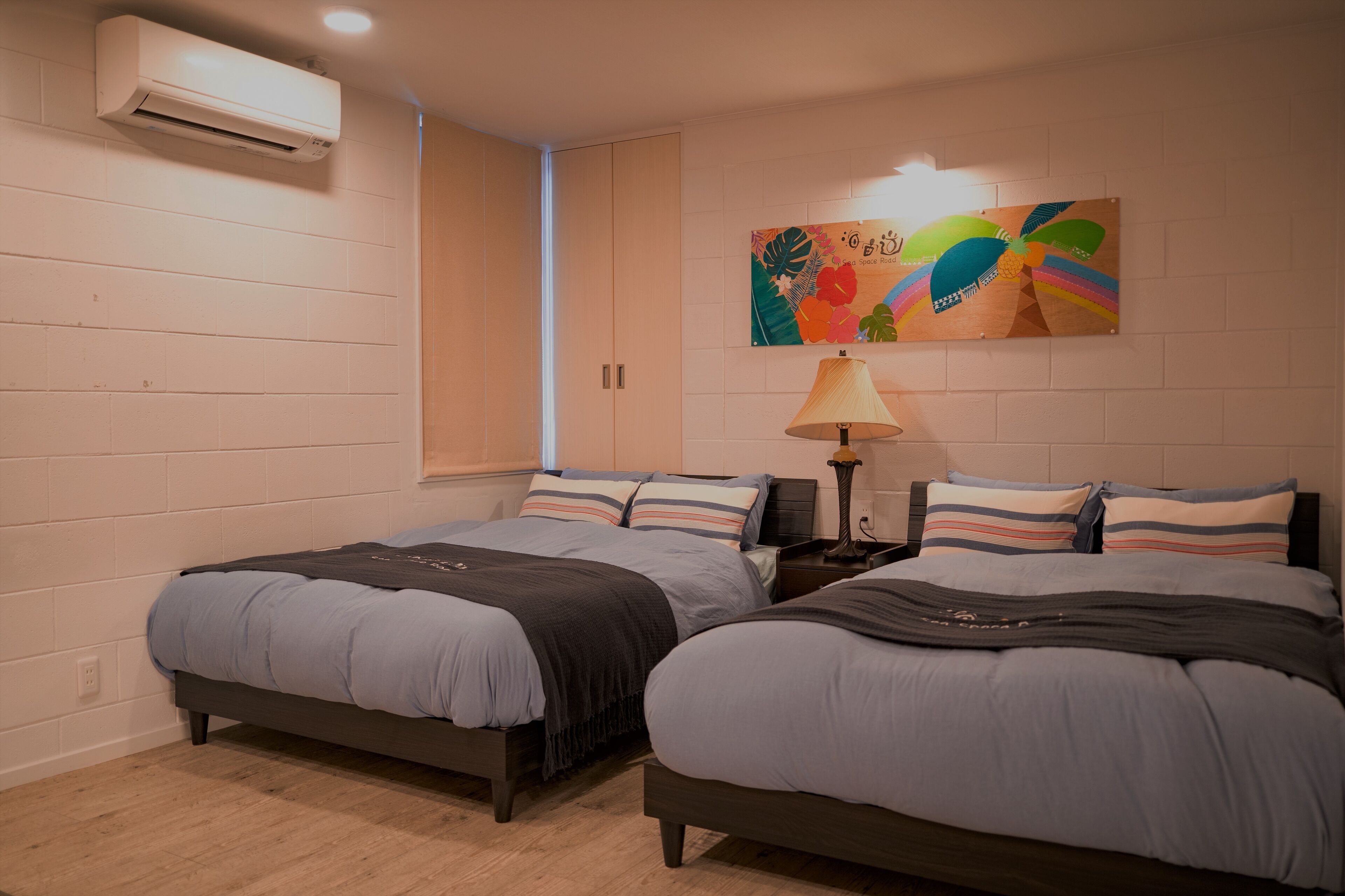 Un dormitorio con dos camas, aire acondicionado de pared, una obra de arte colorida y una lámpara de noche.