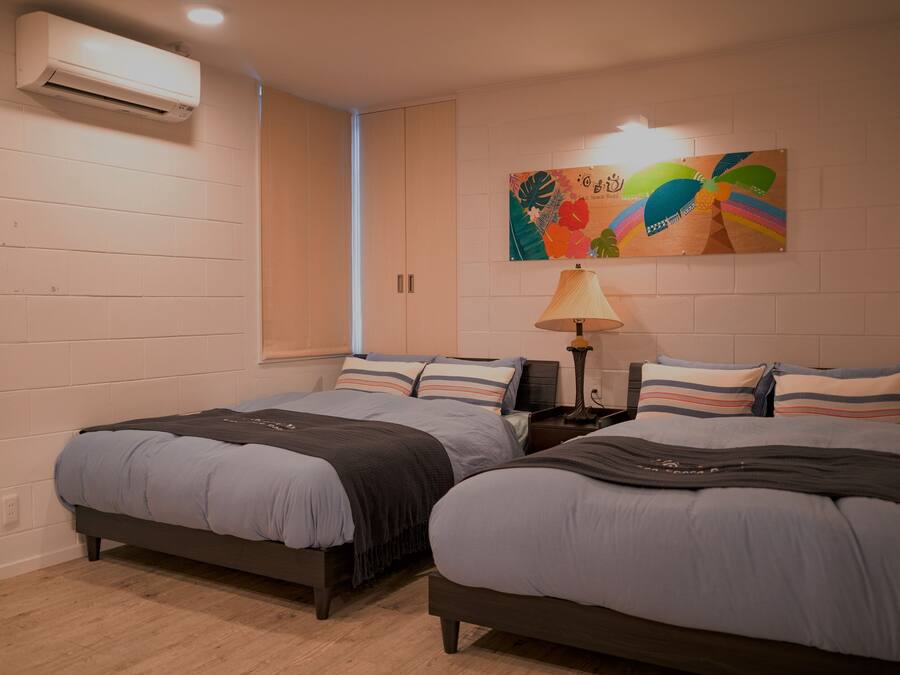 Un dormitorio con dos camas, aire acondicionado de pared, una obra de arte colorida y una lámpara de noche.