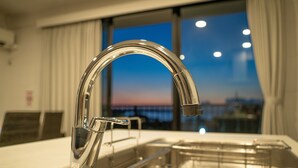A modern kitchen with a stainless steel sink and faucet, a window with a view of the cityscape at dusk, and a clean, minimalist design. - Sea Space Road (Onna)