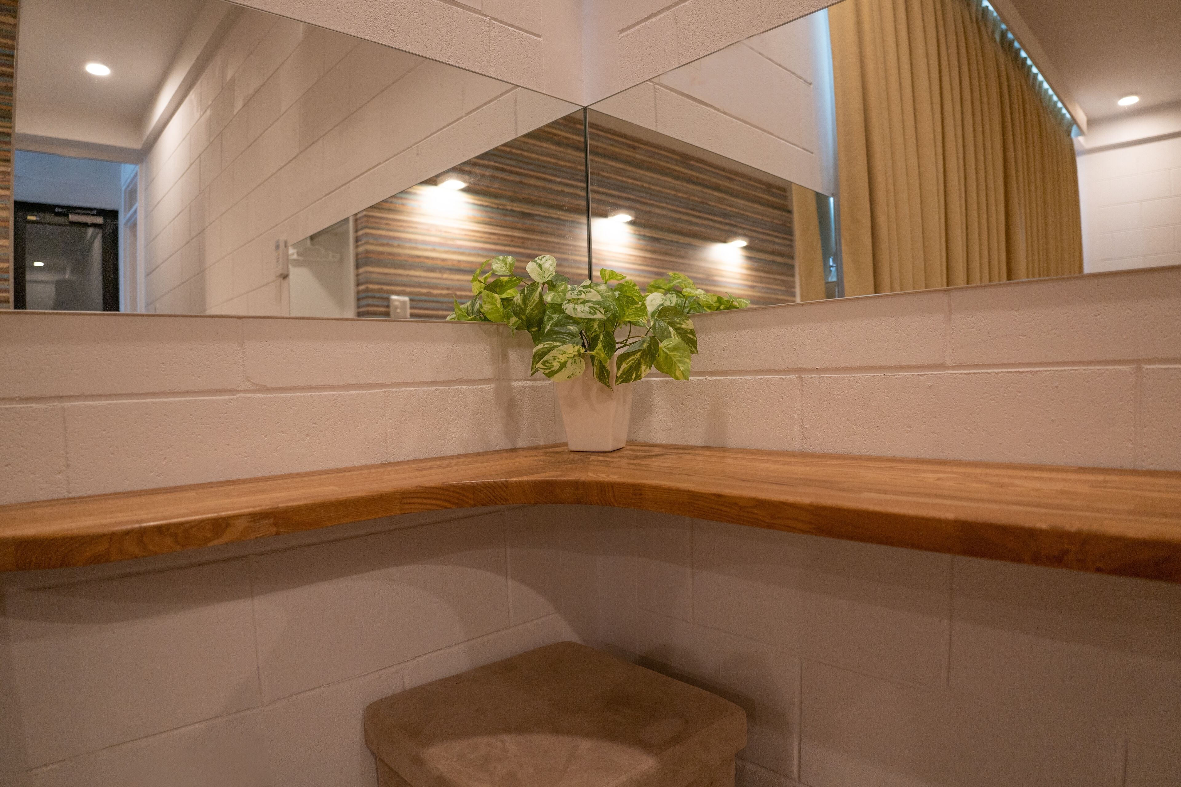 A corner of a hotel room with a wooden bench, a small stool, and a potted plant on a white brick wall.