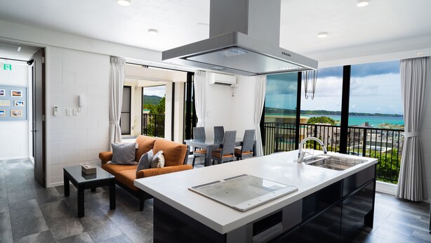 A modern kitchen with a large island, a dining area with a table and chairs, and a balcony with a view of the sea. - Sea Space Road (Onna)