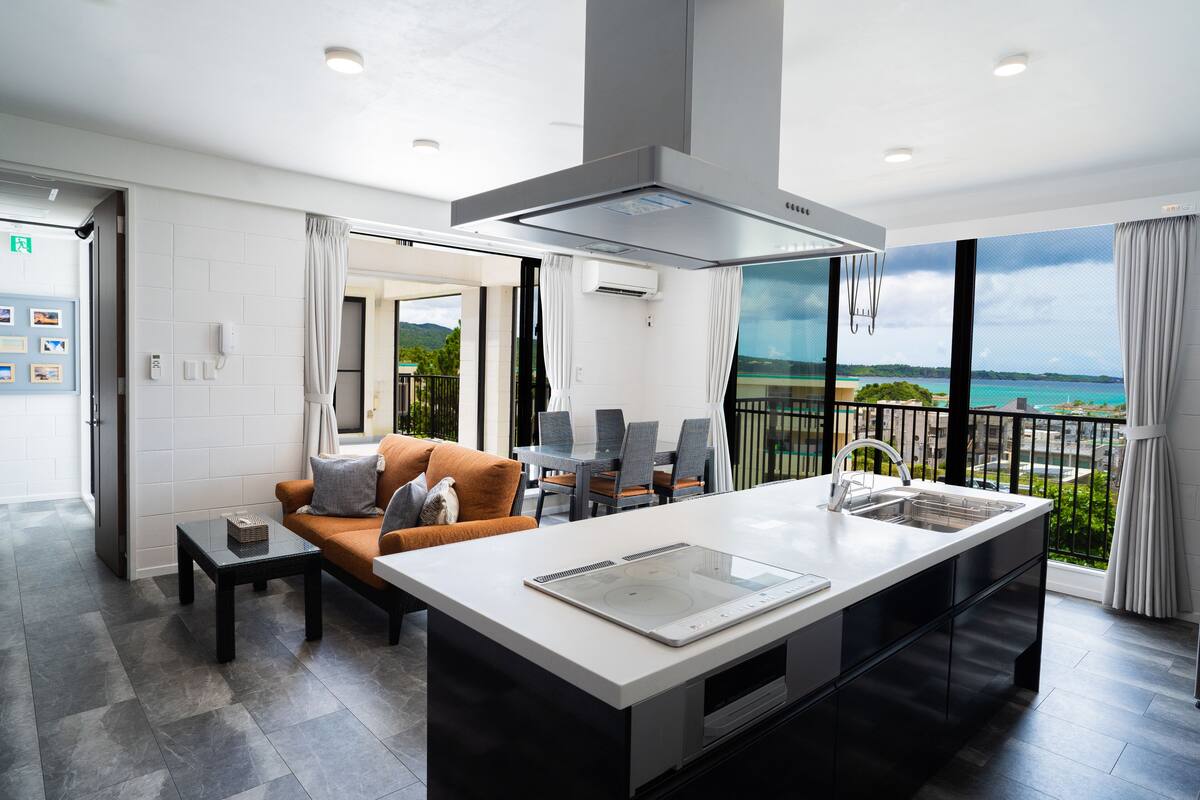 A modern kitchen with a large island, a dining area with a table and chairs, and a balcony with a view of the sea.