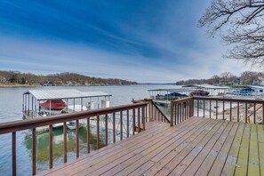 A wooden deck with a railing overlooking a calm body of water, docks with boats, and a distant shoreline with buildings. - Furnished Deck, Dock! Lake of the Ozarks Haven (Sunrise Beach)