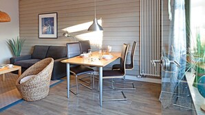 A modern living room with a wooden floor, a dining table with chairs, a sofa, a wicker chair, and a radiator. - Pure bliss Mandö, Germany (List)