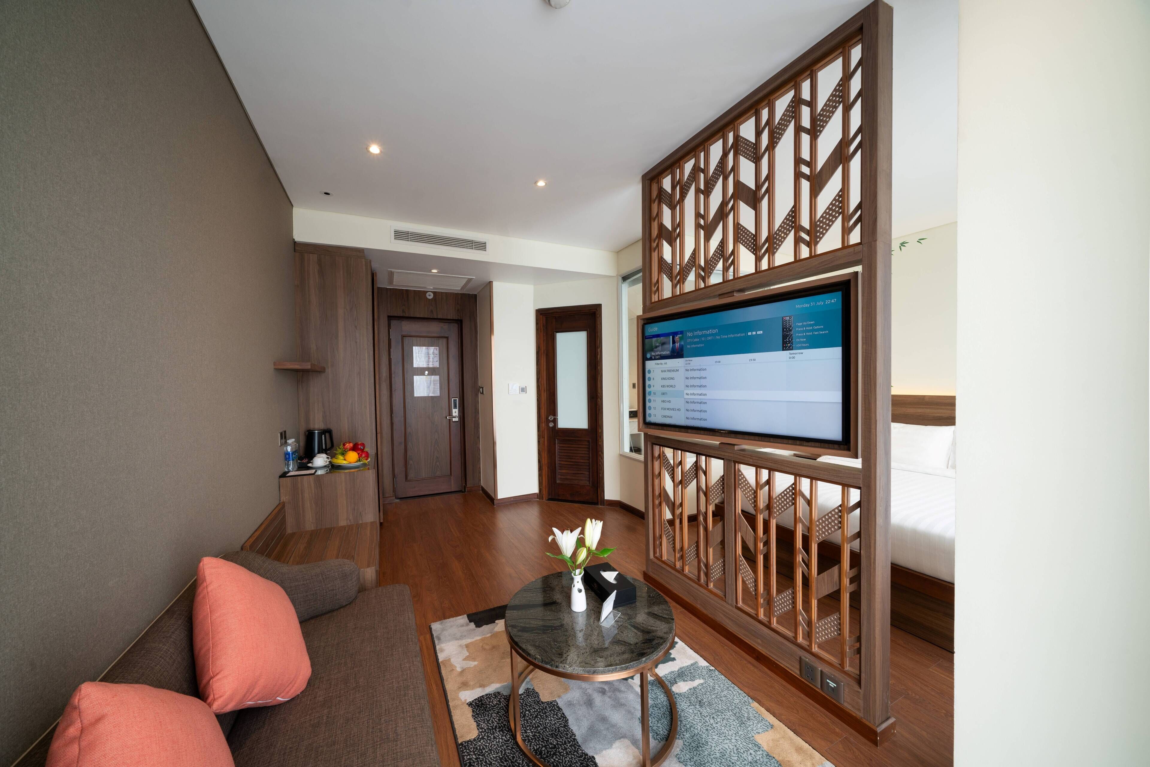 Una sala de estar moderna con un sofá, una mesa de centro y un televisor. Hay un separador de madera con un panel decorativo, y una puerta que conduce a otra habitación.