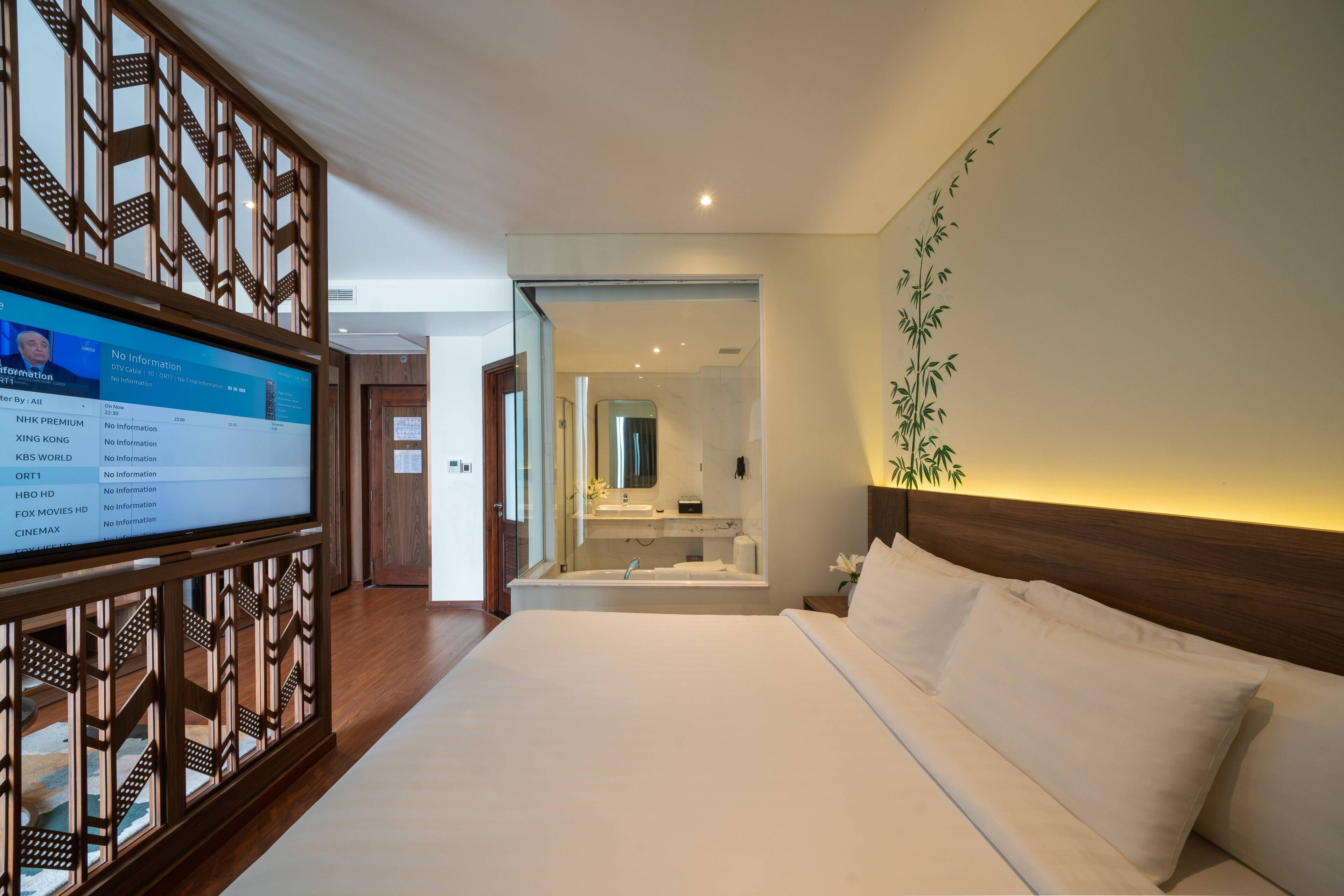 Ein Hotelzimmer mit einem großen Bett, einem an einer Holzvertäfelung montierten Fernseher, einem Badezimmer mit Spiegel und einer Wandpflanze.