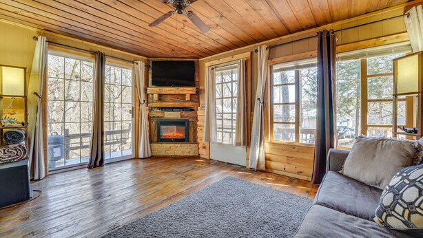 Un interior de cabaña acogedor con chimenea, pisos de madera, ventilador de techo y ventanas amplias.