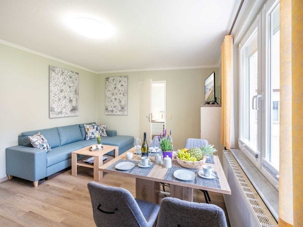 A modern living room with a sofa, dining table, and chairs. There is a TV on a stand and a window with a view.