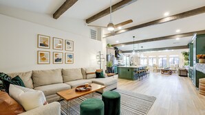 Ein geräumiges Wohnzimmer mit einer Couch, Poufs und einem Couchtisch. Im Hintergrund befindet sich eine Küchenzeile mit Esstisch und Stühlen.