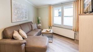A living room with a brown sofa, a coffee table, a radiator, and a window with curtains. - Ahlbeck Villa Stephanie Bungalow H2AS rear - Ahlbeck Villa Stephanie (Heringsdorf)