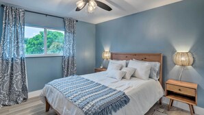 Une chambre à coucher avec un lit, un ventilateur de plafond, une table de chevet avec une lampe et une fenêtre avec des rideaux.