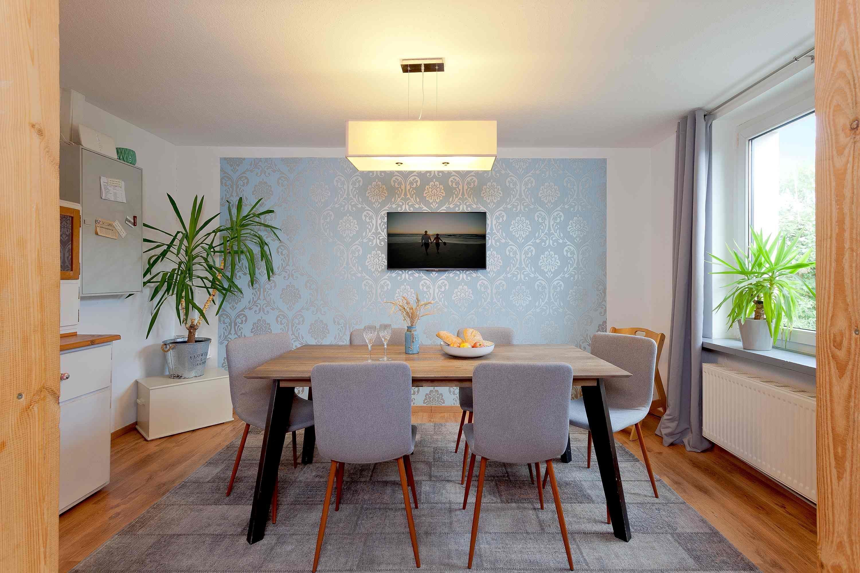 A dining area with a wooden table, four grey chairs, a potted plant, and a framed picture on the wall.