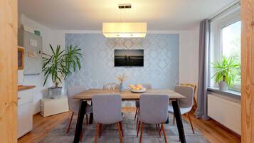 A dining area with a wooden table, four grey chairs, a potted plant, and a framed picture on the wall.