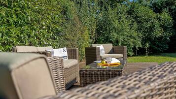 A wicker outdoor seating area with cushioned chairs and a glass-top table, surrounded by lush greenery.