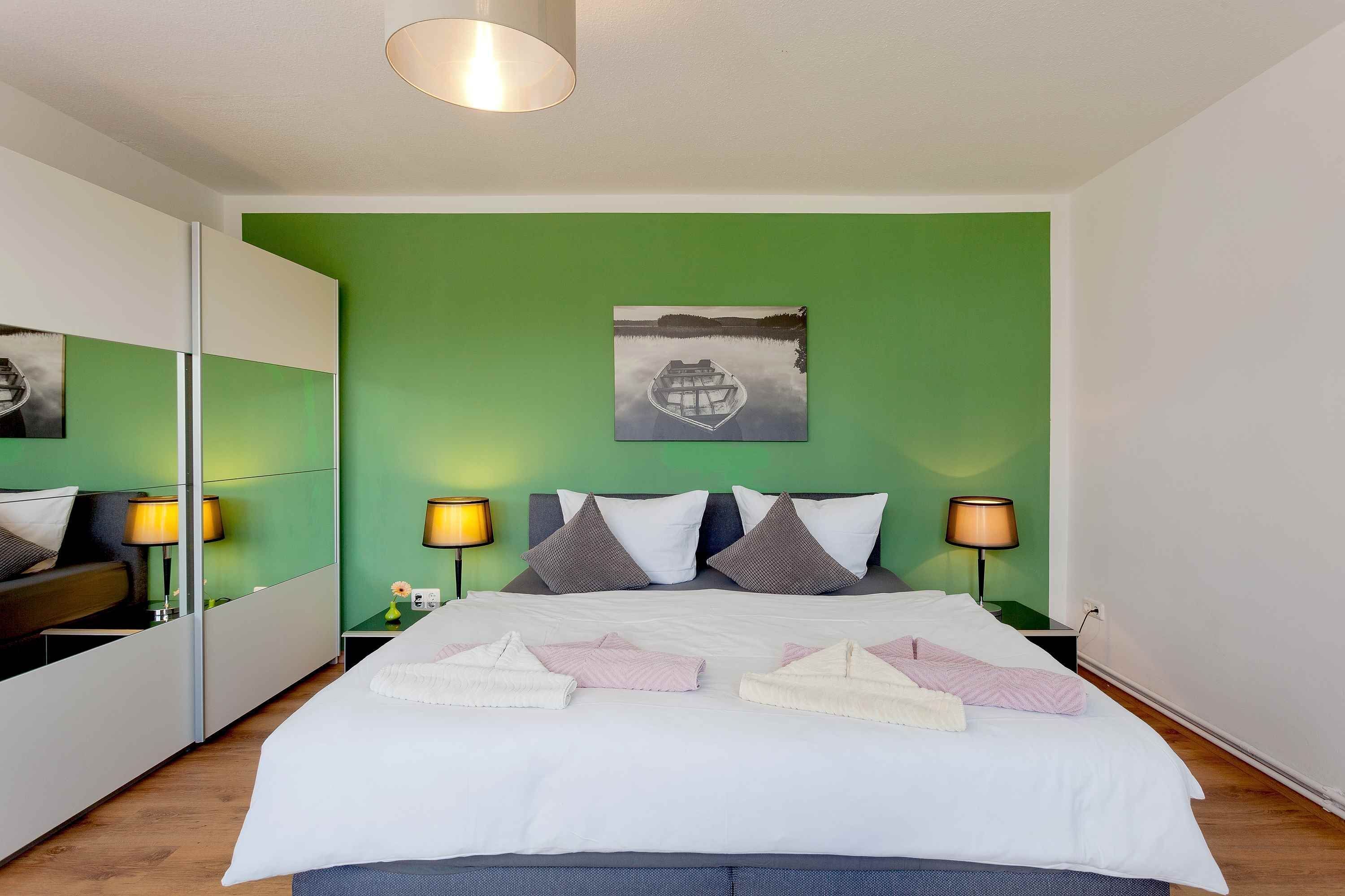 A hotel room with a green wall, a bed with white bedding, two nightstands with lamps, and a painting of a boat on the wall.